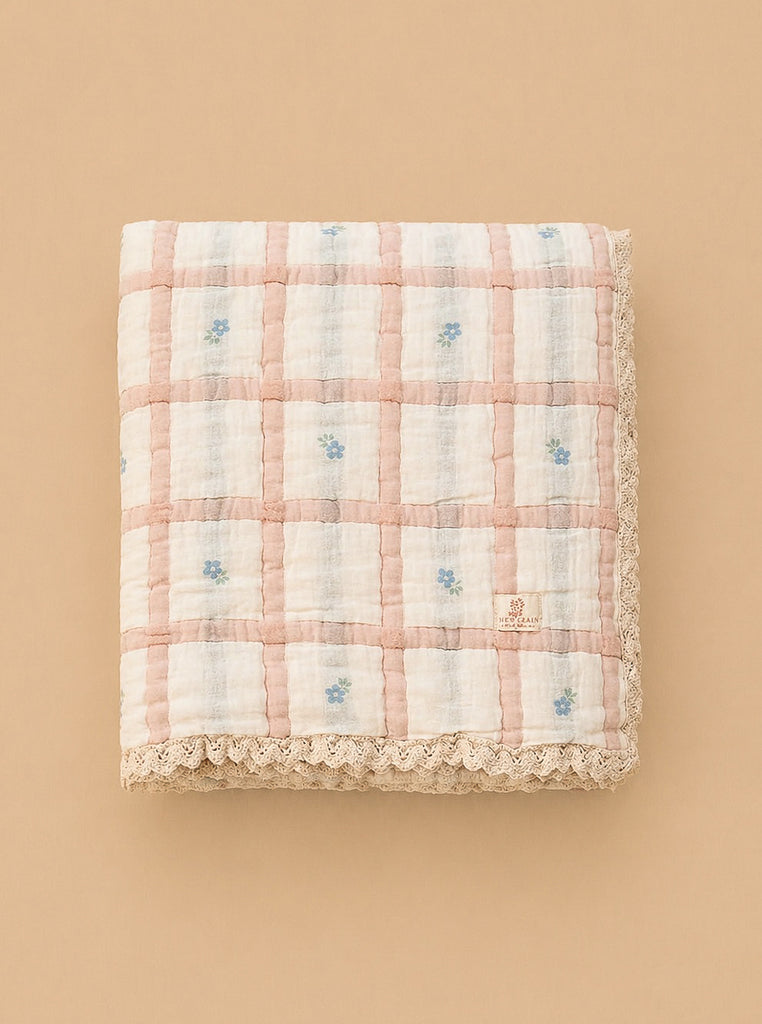 The Organic Cotton Baby Quilt - Meadow, Petal, made in Portugal, features a pink and white checkered design with delicate blue flowers and lace trim, displayed on a neutral beige background.