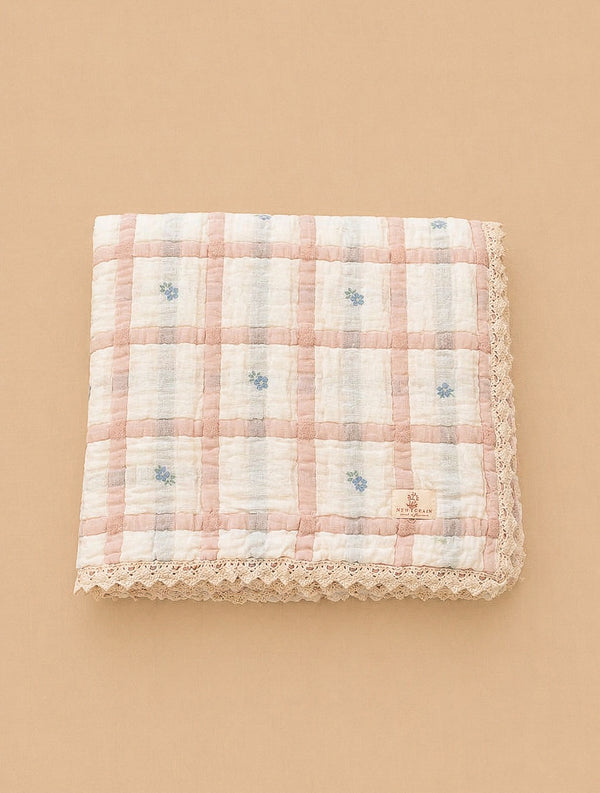The Organic Cotton Twin Size Quilt - Meadow, Petal features a pink and white checkered pattern with small blue floral accents and lace trim, made in Portugal, shown folded on a beige background.