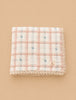 The Organic Cotton Twin Size Quilt - Meadow, Petal features a pink and white checkered pattern with small blue floral accents and lace trim, made in Portugal, shown folded on a beige background.