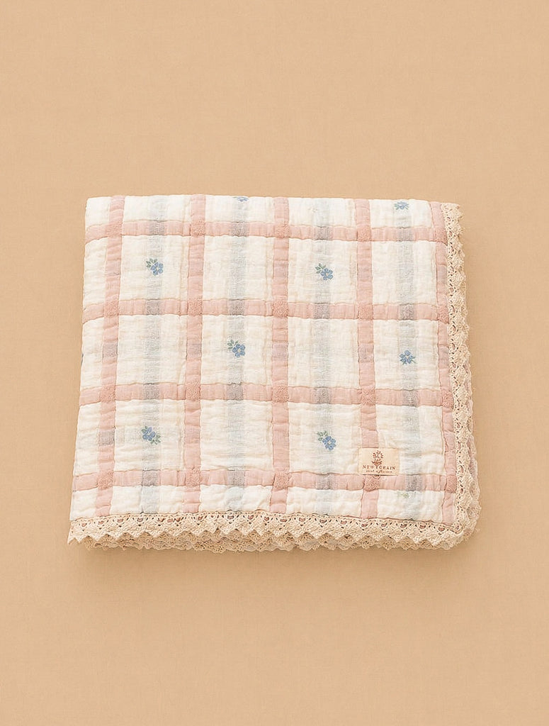 The Organic Cotton Twin Size Quilt - Meadow, Petal features a pink and white checkered pattern with small blue floral accents and lace trim, made in Portugal, shown folded on a beige background.