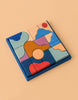 Grimm's Small Building Block Set - Wave features abstract, irregularly shaped wooden pieces in blue, red, pink, orange, and tan—perfect for creative building—arranged within a square blue frame on a beige background.