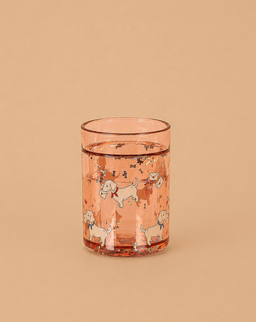 The Konges Sloejd Glitter Cup - Woof Letter Rose (Single) is a pink kids’ cup featuring playful dog, bone, and star designs, filled with water and set against a plain beige background.