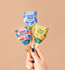 A hand with purple nail polish holds three Garden Seed Pop (Single) sticks—Sweet Basil, Strawberry, and Carrot Bunch—against a beige background.