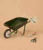 The Heritage Wheelbarrow – Liane, a green toy wheelbarrow with light handles, rests on a tan background with floral gardening gloves over the handles and a kids garden tool set—a small green hand trowel and hand rake—nearby.