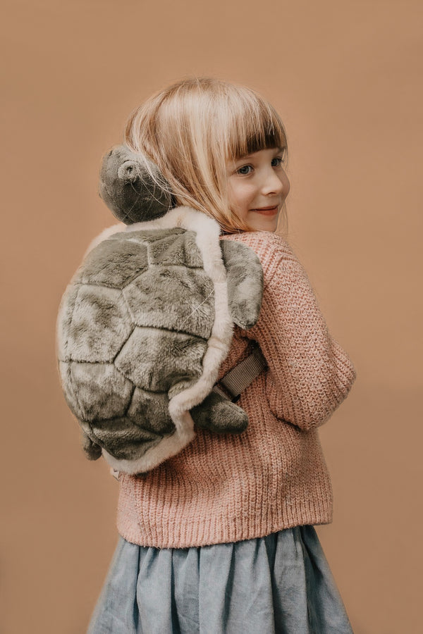 A young child with blonde hair, wearing a pink sweater and blue skirt, smiles over their shoulder while carrying the Children's Backpack - Turtle with adjustable straps against a plain beige background.