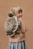 A young child with blonde hair, wearing a pink sweater and blue skirt, smiles over their shoulder while carrying the Children's Backpack - Turtle with adjustable straps against a plain beige background.