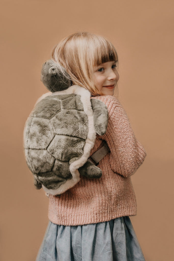 A young child with blonde hair, wearing a pink sweater and blue skirt, smiles over their shoulder while carrying the Children's Backpack - Turtle with adjustable straps against a plain beige background.