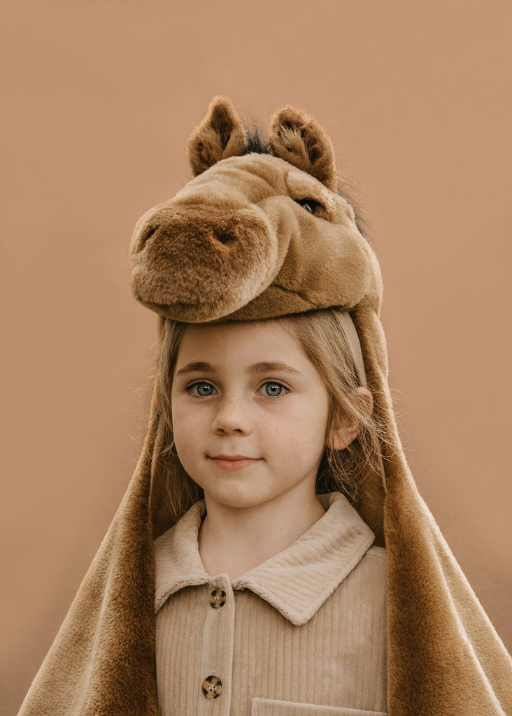 A young child with light hair wears the Costume - Brown Horse, featuring a plush brown horse hat with ears and mane, paired with a beige button-up shirt, standing against a plain tan background.