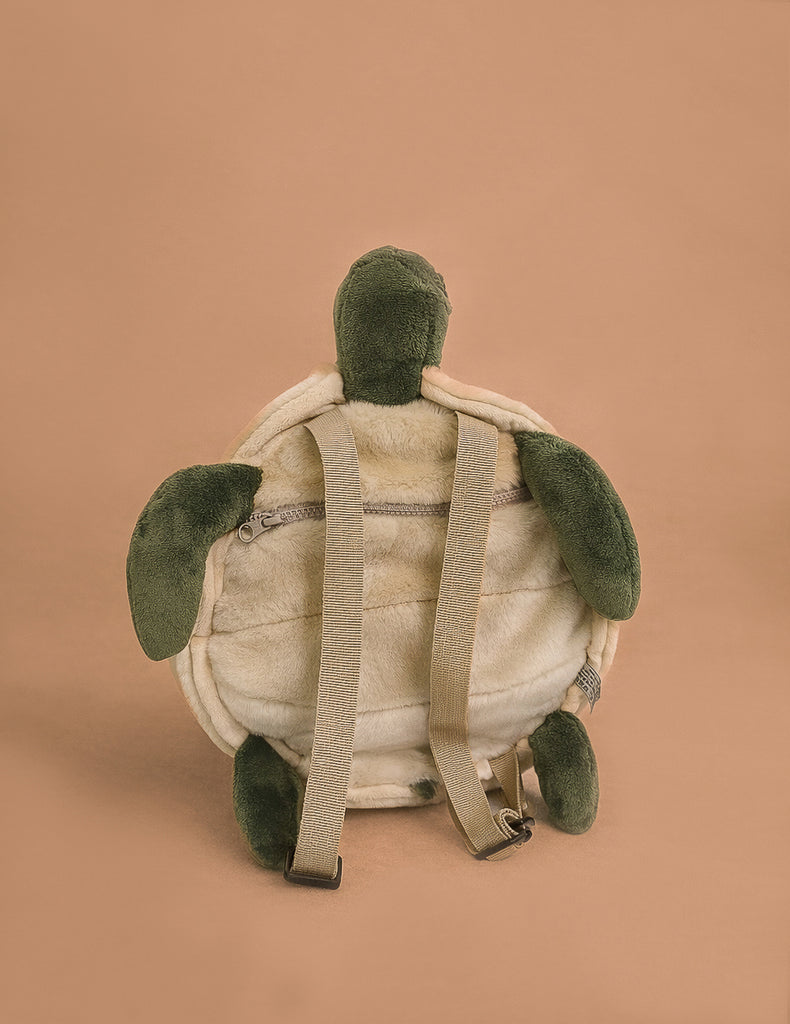 The Children's Backpack - Turtle features green flippers and head, a beige shell, adjustable straps, and is displayed on a tan background with its back and zipper showing.
