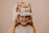 A smiling blonde-haired child wears the Costume - Tiger, a large plush faux fur hat with ears and face, paired with a cream knit sweater, standing against a plain beige background.