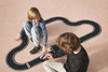 Two children play on a light-colored floor with the Large Flexible Toy Race Track - Grand Prix. One child drives a blue car along the flexible track as the other watches, both looking engaged and happy.