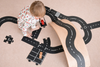 A toddler in pajamas plays on the floor with toy cars and a wooden ramp, arranging black road pieces from the WaytoPlay: Extra Long Flexible Road Set - King of the Road, complete with white markings, to create a miniature roadway scene. The cityscape construction sparks imaginative play as new routes and adventures unfold.