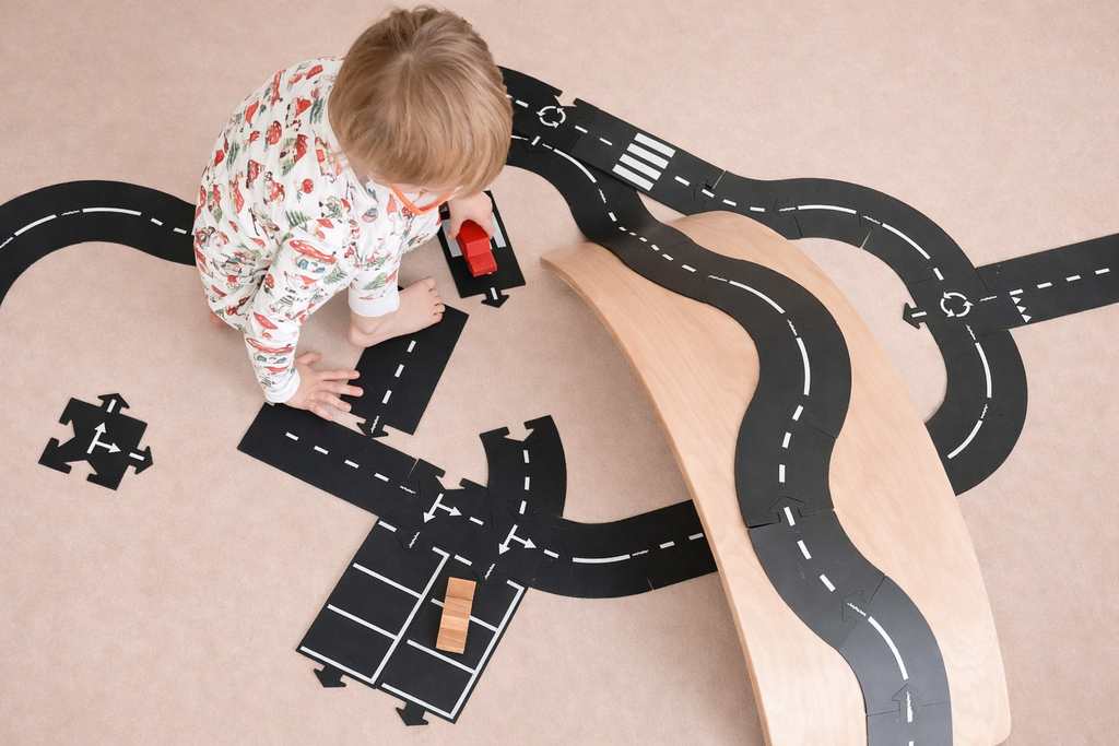 A toddler in pajamas plays on the floor with toy cars and a wooden ramp, arranging black road pieces from the WaytoPlay: Extra Long Flexible Road Set - King of the Road, complete with white markings, to create a miniature roadway scene. The cityscape construction sparks imaginative play as new routes and adventures unfold.