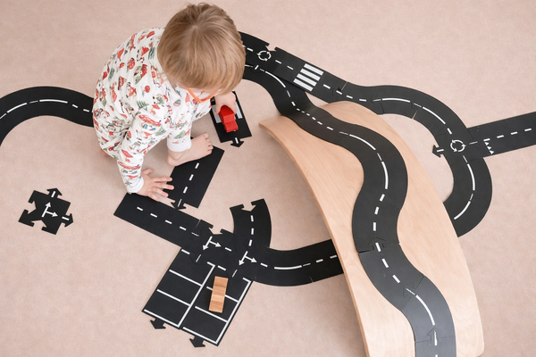 A toddler in pajamas plays on the floor with toy cars and a wooden ramp, arranging black road pieces from the WaytoPlay: Extra Long Flexible Road Set - King of the Road, complete with white markings, to create a miniature roadway scene. The cityscape construction sparks imaginative play as new routes and adventures unfold.