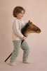 A young child with curly hair, dressed in a beige sweater and light green pants, smiles as they ride the Pretend Play Ride-On Horse, sparking imaginative adventures against a neutral background.