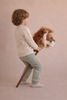 A young child with light brown hair, dressed in a cream sweater and light green pants, holds the Hobbyhorse - Lion toy and stands sideways, ready for adventure against a plain beige background.