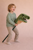 A young child in a green sweater and beige pants smiles while riding the Hobbyhorse - T-Rex Dino, a stick toy with a plush dinosaur head, enjoying imaginative play against a neutral background.