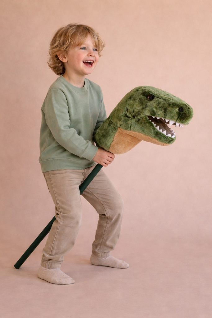 A young child in a green sweater and beige pants smiles while riding the Hobbyhorse - T-Rex Dino, a stick toy with a plush dinosaur head, enjoying imaginative play against a neutral background.