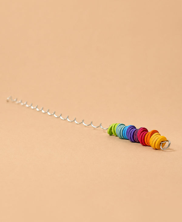 A Grimm's Spirelli Clatter Wand featuring a silver spiral stick with vibrant, round beads stacked in rainbow order at one end, reminiscent of a classic wooden spiral toy, set against a plain beige background.