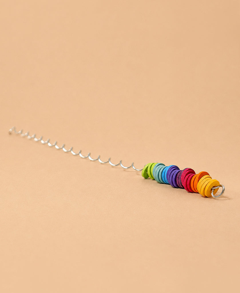 A Grimm's Spirelli Clatter Wand featuring a silver spiral stick with vibrant, round beads stacked in rainbow order at one end, reminiscent of a classic wooden spiral toy, set against a plain beige background.