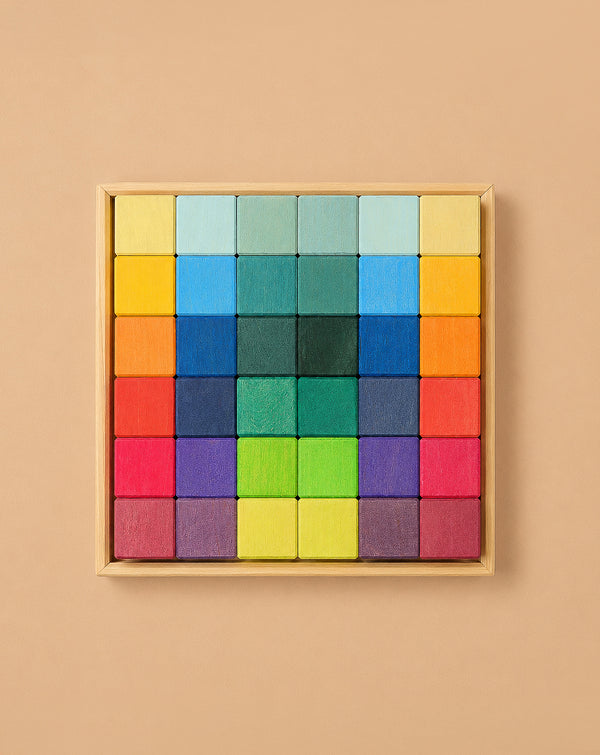 Grimm's Rainbow Mosaic Building Blocks are neatly arranged in a wooden tray, featuring a symmetrical, gradient pattern that transitions from warm reds, oranges, and yellows along the edges to cool greens, blues, and purples at the center—ideal for promoting fine motor skills and spatial thinking through engaging play.