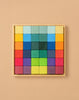 Grimm's Rainbow Mosaic Building Blocks are neatly arranged in a wooden tray, featuring a symmetrical, gradient pattern that transitions from warm reds, oranges, and yellows along the edges to cool greens, blues, and purples at the center—ideal for promoting fine motor skills and spatial thinking through engaging play.