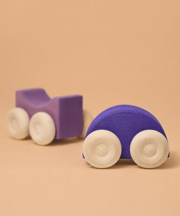 Two Grimm's Stacking Cars - Purple with natural-colored wheels sit on a light brown surface: a rounded purple car in front and a lavender square-shaped car behind, making these charming wooden cars ideal educational toys.