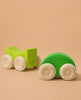 Two green wooden cars with white wheels rest on a light brown surface. The closer car is rounded, while the other, set further back, is blockier and inspired by Grimm's Stacking Cars - Green.