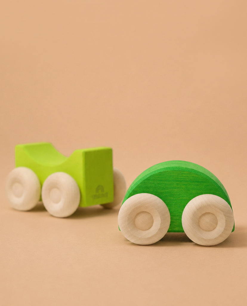 Two green wooden cars with white wheels rest on a light brown surface. The closer car is rounded, while the other, set further back, is blockier and inspired by Grimm's Stacking Cars - Green.