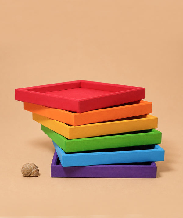 Six Grimm’s Rainbow Frames trays in vivid red, orange, yellow, green, blue, and purple are stacked in a slanted tower on a beige background. A snail shell sits beside them—ideal for brightening any kid’s workspace.