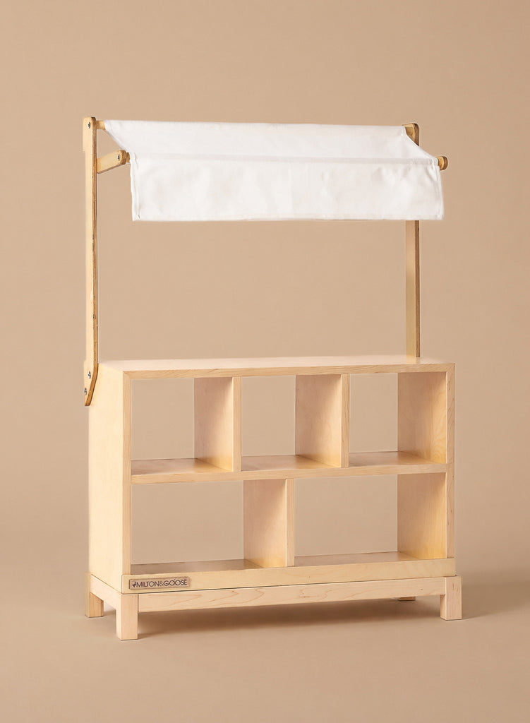 The Milton & Goose Market Stand, handcrafted in the USA, features six open cubby shelves and a white fabric canopy—ideal for pretend play—shown here against a plain beige background.