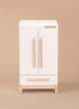 The Milton & Goose Essential Play Refrigerator—made in the USA—is a white wooden cabinet with two vertical handles, a lower drawer, and short natural wood legs, ideal for storing play food and kitchen toys on a plain beige background.
