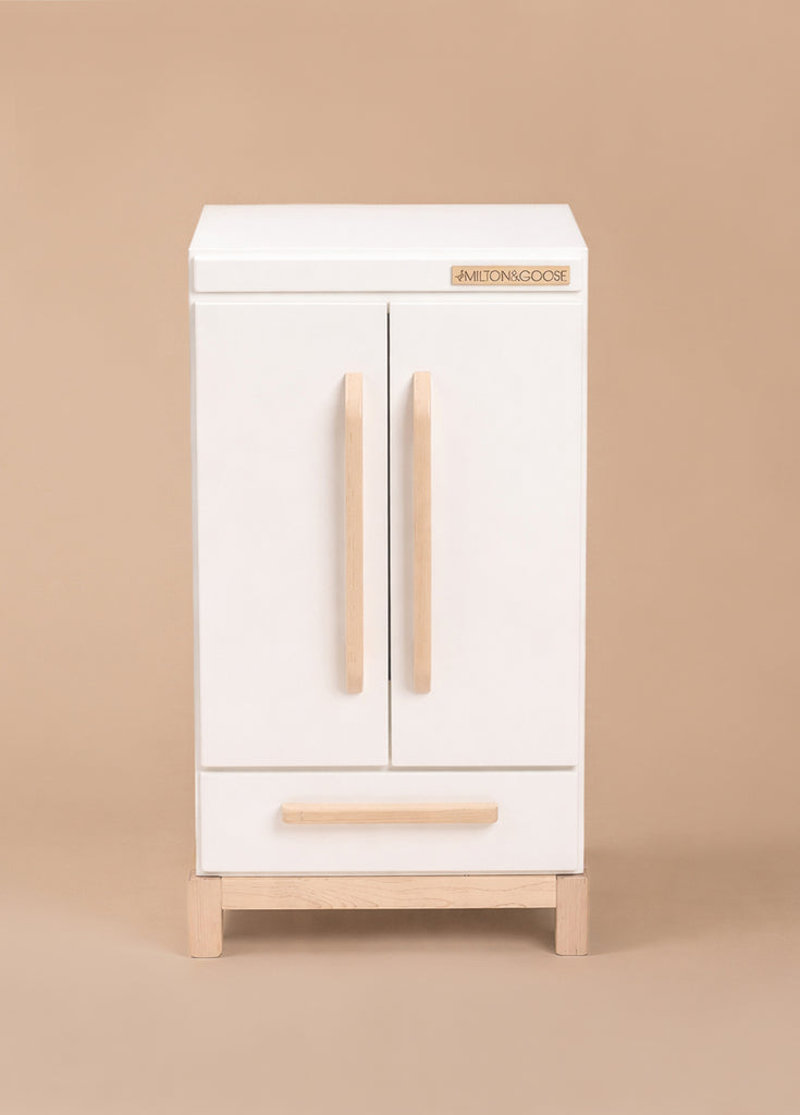The Milton & Goose Essential Play Refrigerator—made in the USA—is a white wooden cabinet with two vertical handles, a lower drawer, and short natural wood legs, ideal for storing play food and kitchen toys on a plain beige background.