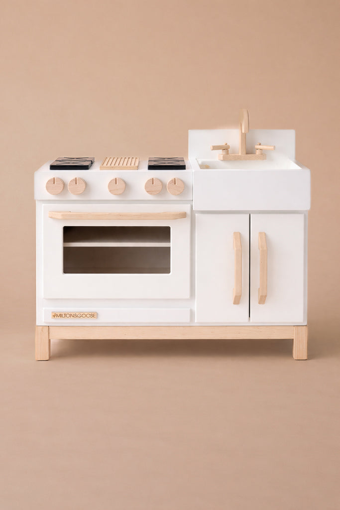 The Milton & Goose Essential Play Kitchen—made in the USA—features a white stove with four burners, an oven with a clear window, a sink and faucet, and wooden handles. Perfect for imaginative play against a beige background.