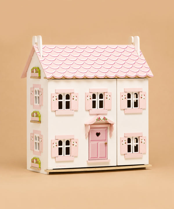Sophie's Wooden Dolls House features a pink roof, white walls, and pink window shutters. Made from FSC-certified wood, it has a pink door with a heart-shaped window, green flower boxes, and sits against a solid beige background.