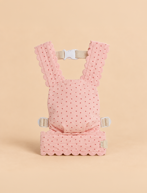The Olli Ella Petal Doll Carrier - Strawberry is a pink baby carrier featuring scalloped edges and small red polka dots. Displayed upright on a beige background, it has white buckles and straps at the top and sides—perfect for carrying your favorite doll.