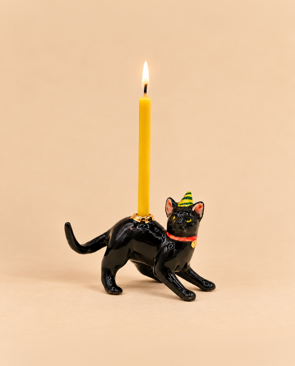 Introducing the Black Cat Cake Topper - Final Sale: a hand-painted collectible featuring a black cat in a festive party hat, carrying a lit yellow candle on its back against a simple beige background. Perfect as a whimsical keepsake birthday gift or unique porcelain cake topper, this charming figurine brings playful delight to any celebration.