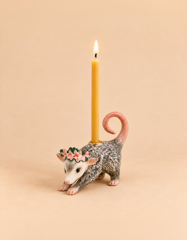 The “Possum Cake Topper - Final Sale” is a hand-painted ceramic possum figurine adorned with a delicate flower crown, carrying a lit yellow candle on its back. Set against a beige background, this charming porcelain piece makes an ideal cake topper and collectible keepsake.