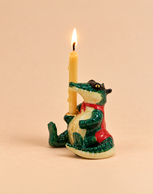 The Later Gator Cake Topper features a handcrafted ceramic crocodile figurine in a red vest holding an upright, lit yellow candle with melted wax dripping down; set against a plain beige background, this adorable collectible is the perfect keepsake or birthday decoration to brighten any celebration.