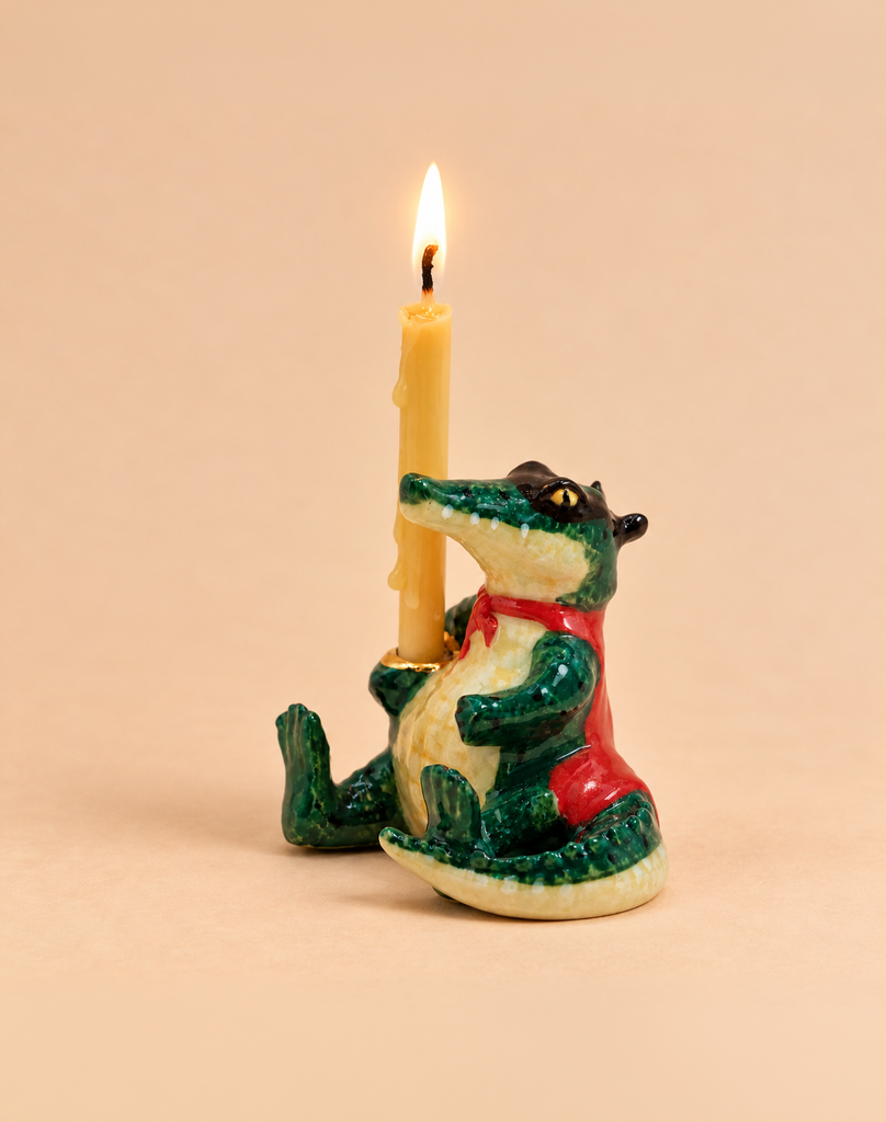 The Later Gator Cake Topper features a handcrafted ceramic crocodile figurine in a red vest holding an upright, lit yellow candle with melted wax dripping down; set against a plain beige background, this adorable collectible is the perfect keepsake or birthday decoration to brighten any celebration.