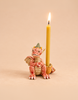 A yellow beeswax birthday candle is lit and held by the Dragon Cake Topper—a red and cream ceramic dragon candle holder with a green hat—on a beige background.