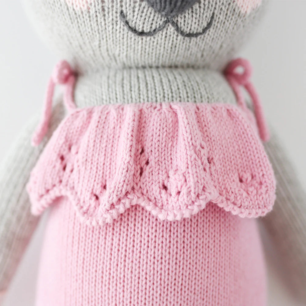 A close-up of Cuddle + Kind Claire the Koala, a hand-knit doll in a pink scalloped dress. Made with light gray yarn and detailed stitching, it highlights the signature craftsmanship. Only her body and part of her face are visible.