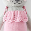 A close-up of Cuddle + Kind Claire the Koala, a hand-knit doll in a pink scalloped dress. Made with light gray yarn and detailed stitching, it highlights the signature craftsmanship. Only her body and part of her face are visible.