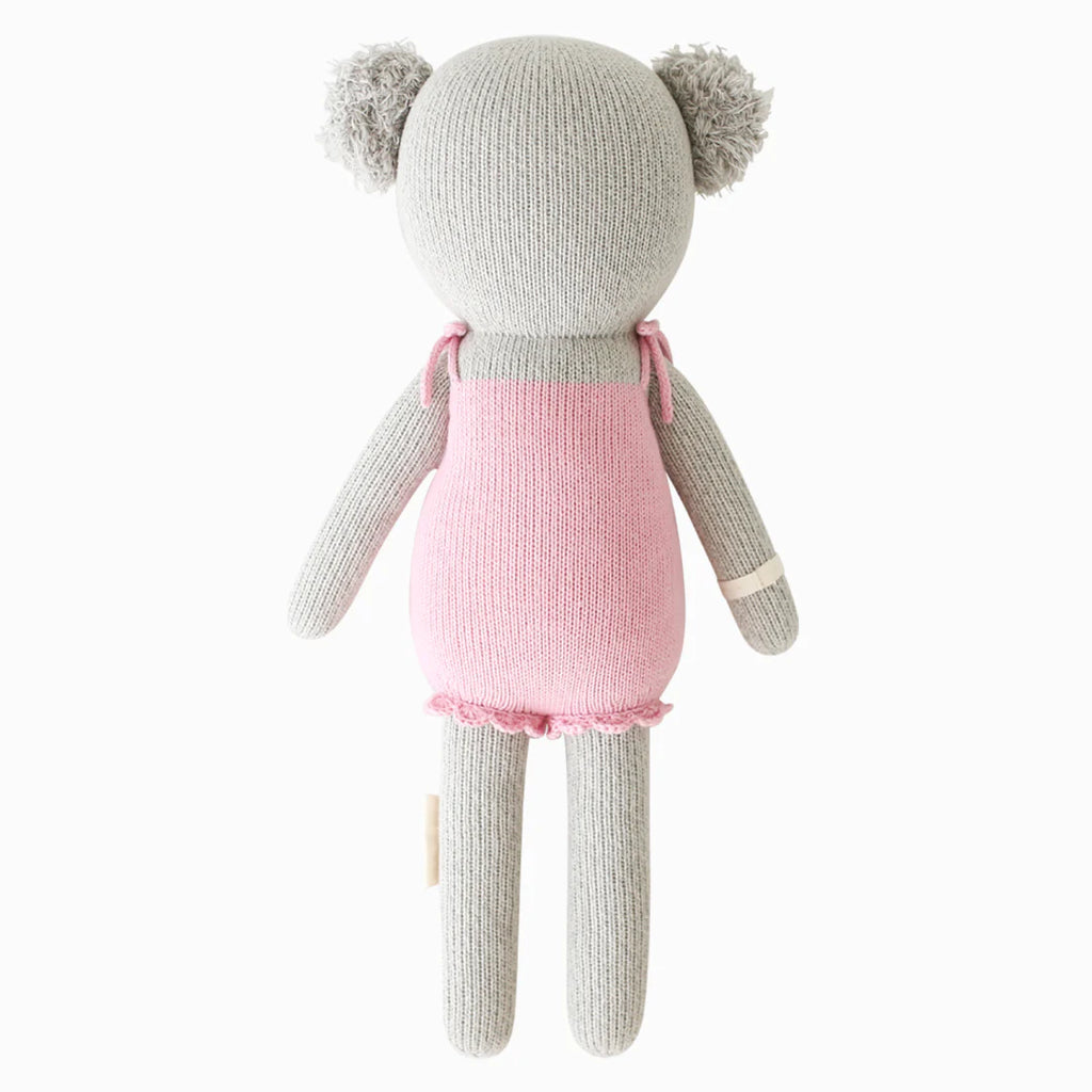 Cuddle + Kind Claire the Koala is a hand-knit grey stuffed animal with round ears, wearing a pink dress and shown from the back against a plain white background.