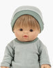 A doll with short brown hair, round cheeks, and a slightly open mouth. The doll is wearing a matching light green beanie and sweater made from soft fabric, reminiscent of Minikane Doll Clothing | Green Tea Fleece Hat. The background is plain white.