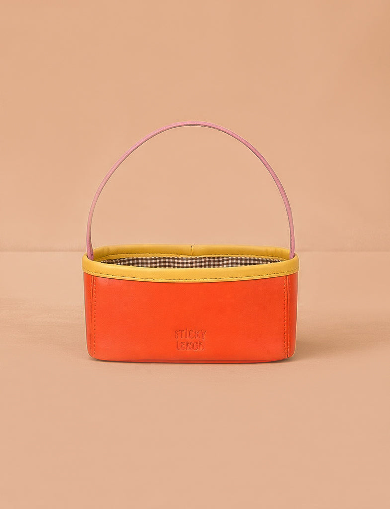 The Sticky Lemon Treasure Bag - Campfire Orange features a rectangular shape with an orange body, yellow trim, pink handle, and checked interior. "STICKY LEMON" is printed on the front—ideal as a playful treasure bag for kids.