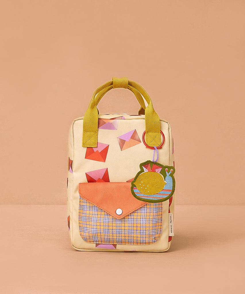The Sticky Lemon Backpack Small - Special Edition - Snail Mail is a small beige kids backpack featuring red and pink geometric shapes, a yellow handle, orange plaid front pocket, green fruit-shaped keychain, and waterproof lining from recycled PET bottles.