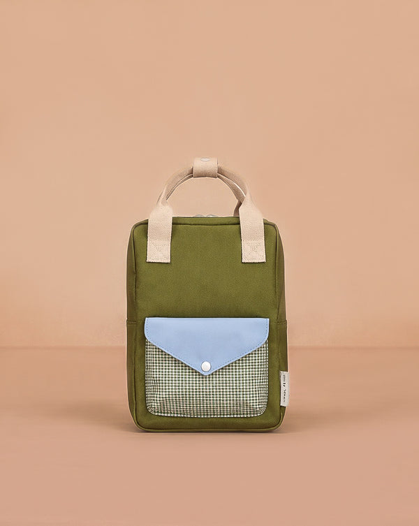 The Sticky Lemon Backpack Small - Envelope - Tickling Grass is a kids’ olive green backpack with beige handles, a blue flap pocket, and a checkered front pocket made from recycled PET bottles, shown on a plain peach background.