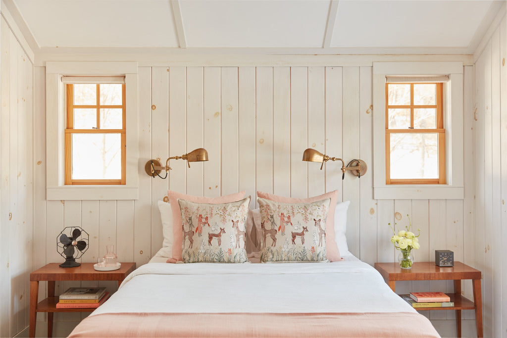 Cozy bedroom with a white-paneled wall, queen size bed with Coral & Tusk Spring Blossoms Pillow and embroidered pillow, brass wall lamps, a small fan, and flowers on the nightstand, bathed in