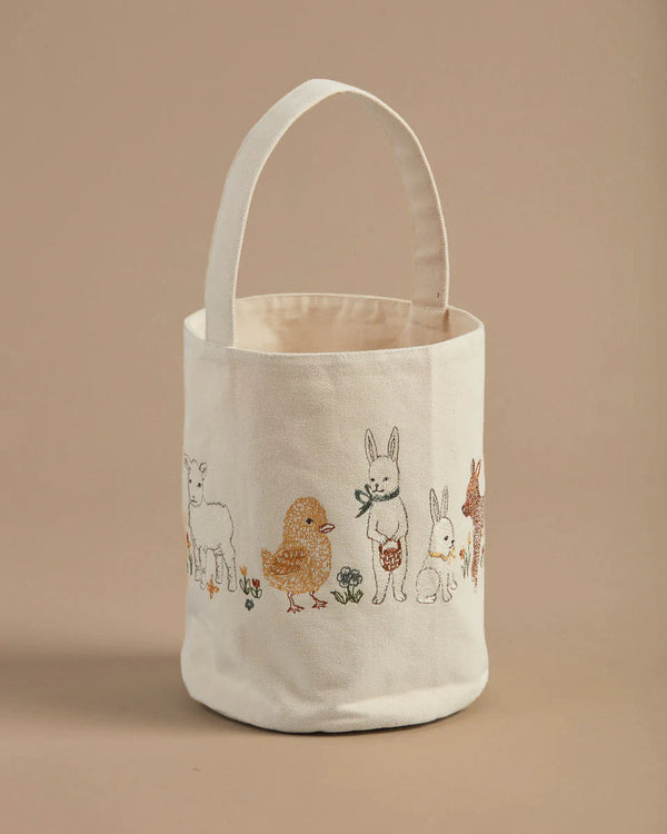The Coral & Tusk Spring Babies Canvas Bucket is a beige fabric tote with a single handle, designed like an Easter basket. It features embroidered images of a lamb, chick, rabbit, other animals, and small plants on a plain light brown background.